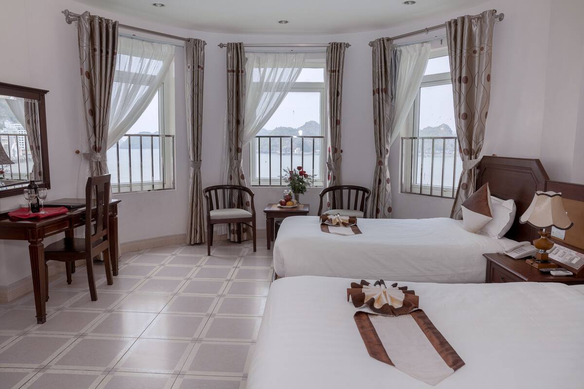 A hotel room with two beds, a desk with a chair, a small table with a vase of flowers, and a view of the sea through large windows.