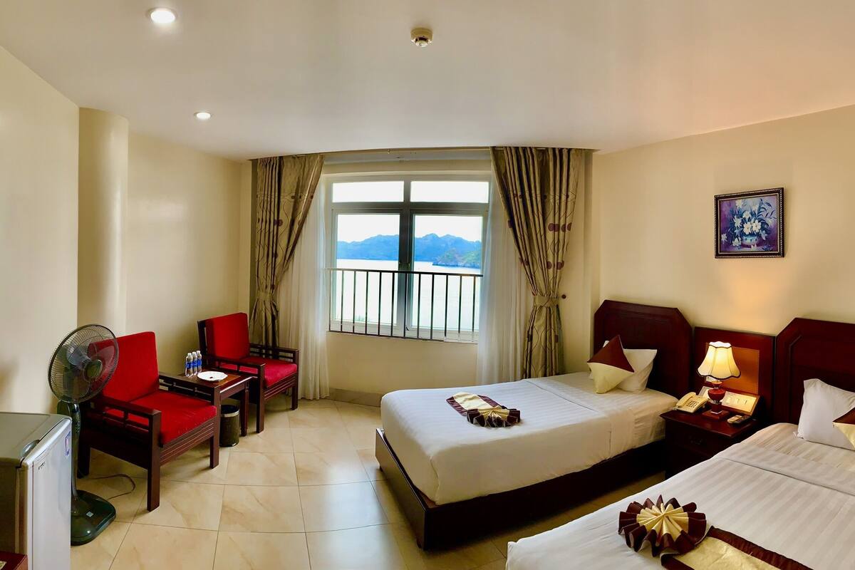 A hotel room with a bed, a desk with a chair, a fan, a window with a view of mountains, and a painting on the wall.