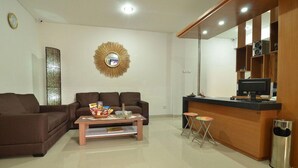 A modern living room with a brown sofa, a coffee table with snacks, a TV, and a small bar area with stools. - Griya Sunset Kuta (Kuta)