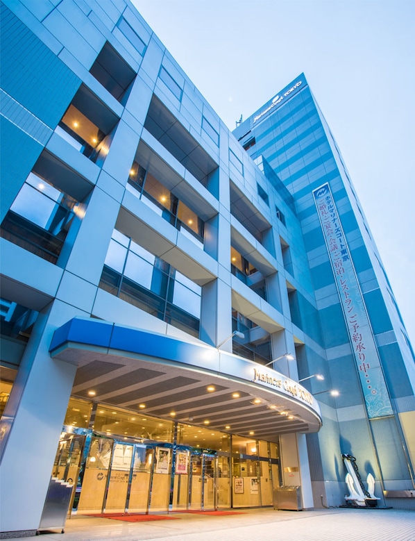 Entrada moderna de hotel com fachada de vidro, área de acesso coberta e placa indicando ser um hotel.