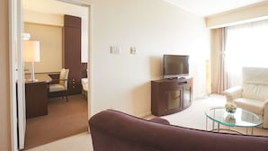 A hotel room with a television, a sofa, a glass coffee table, a desk with a lamp, and a bed.