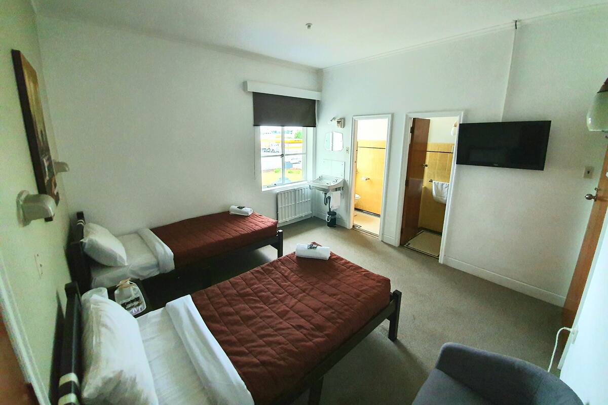 A room with two single beds, a television mounted on the wall, and a window with a view of the outside.