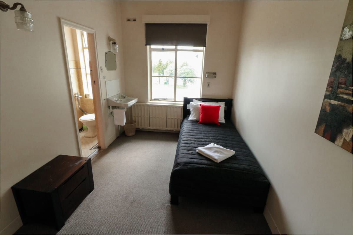 A small, narrow hotel room with a single bed, a window with a view, a radiator, and a small wooden cabinet.