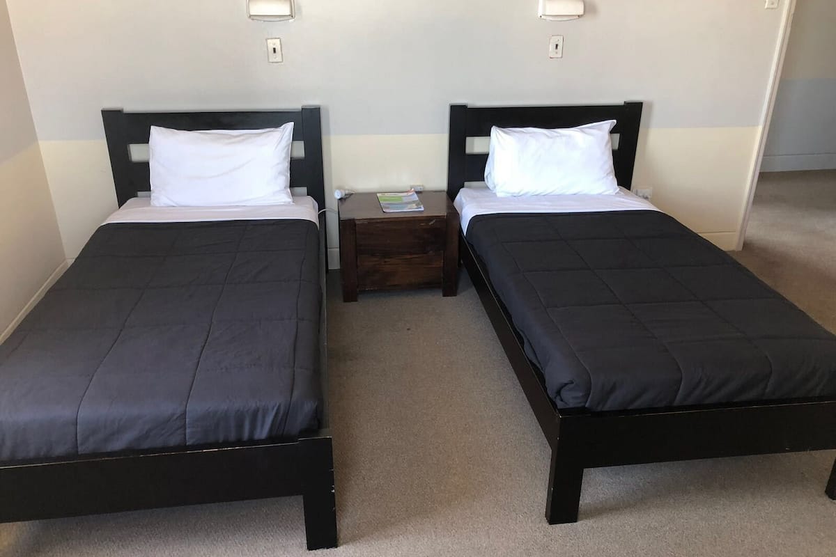 Two single beds with black frames and white bedding, a small wooden nightstand between them, and a light switch on the wall.