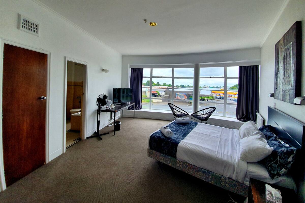 A hotel room with a bed, a desk with a computer, a TV, and a large window with a view of the outside.