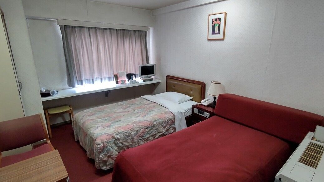 Ein Hotelzimmer mit zwei Betten, einem Schreibtisch mit Computer, einem Stuhl und einem Fenster mit Vorhängen.