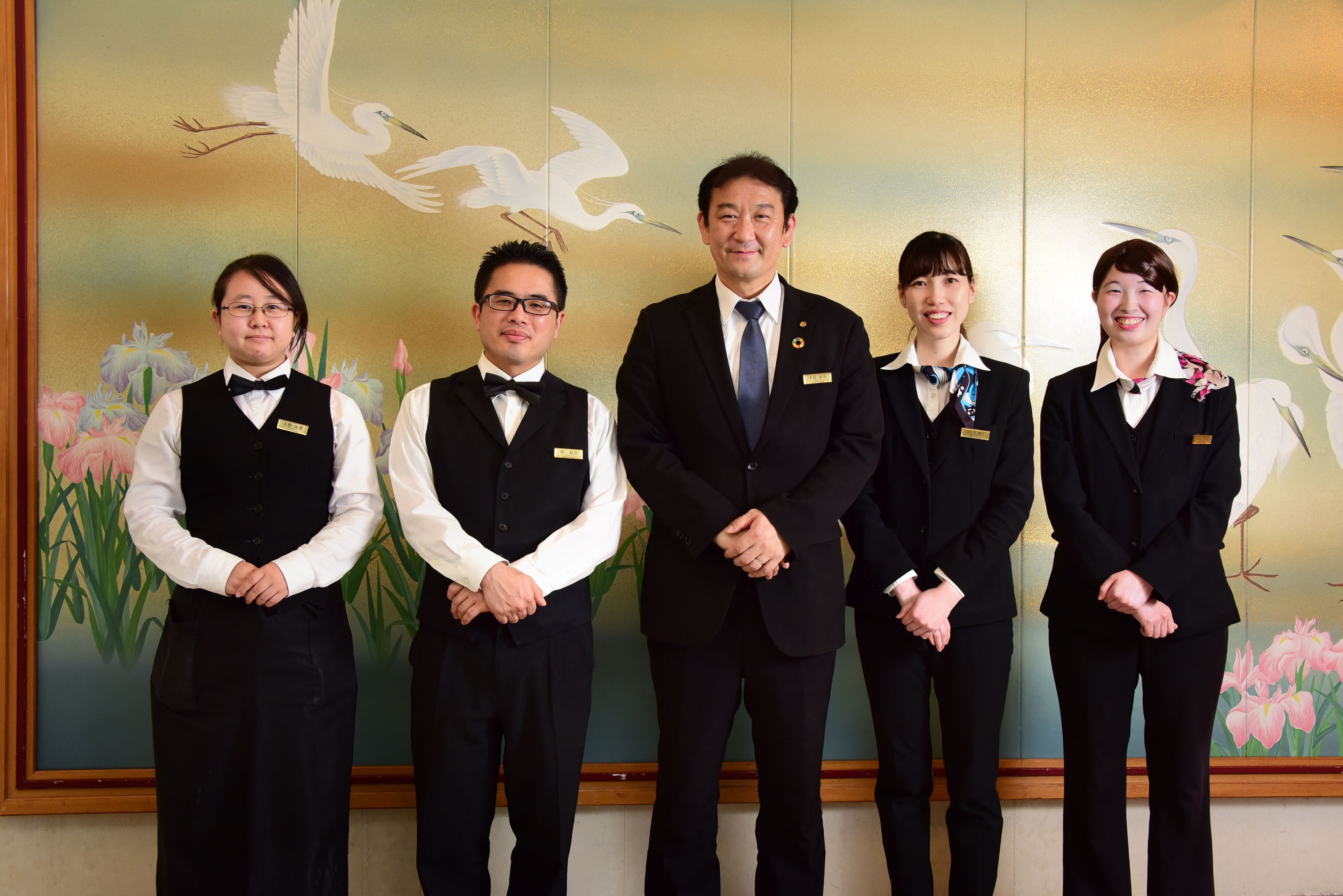 A group of five individuals in formal attire standing in front of a wall with a painting of birds and flowers.