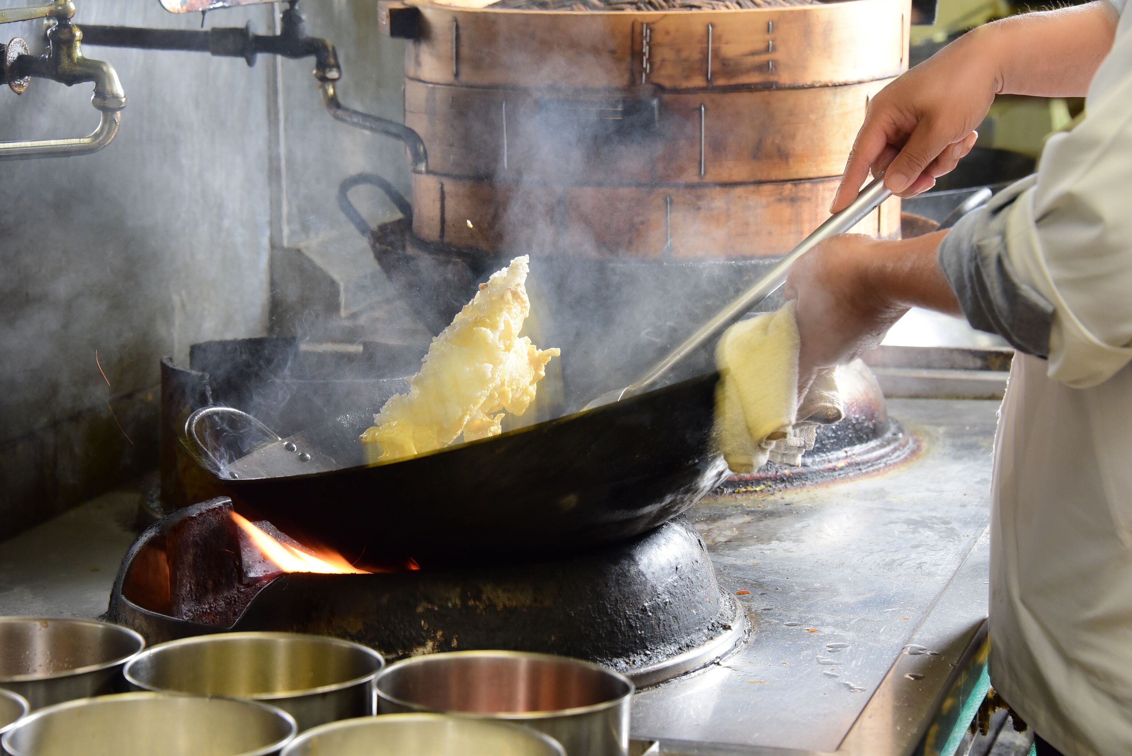 A person cooking in a wok on a stove, with steam rising and several metal bowls on the counter.