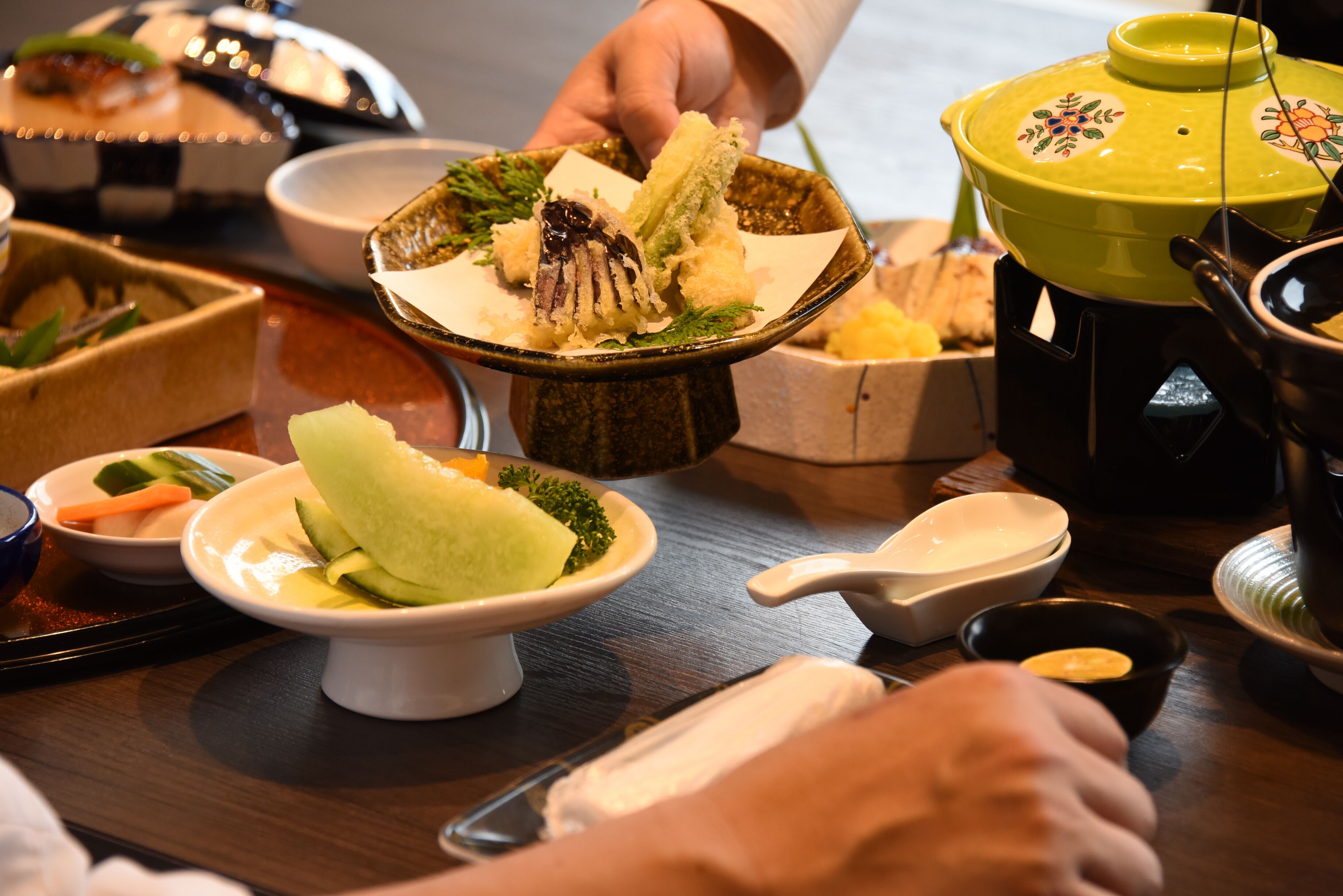 A dining table with various dishes, including a bowl with a green vegetable and a piece of meat, and a yellow pot with a lid.