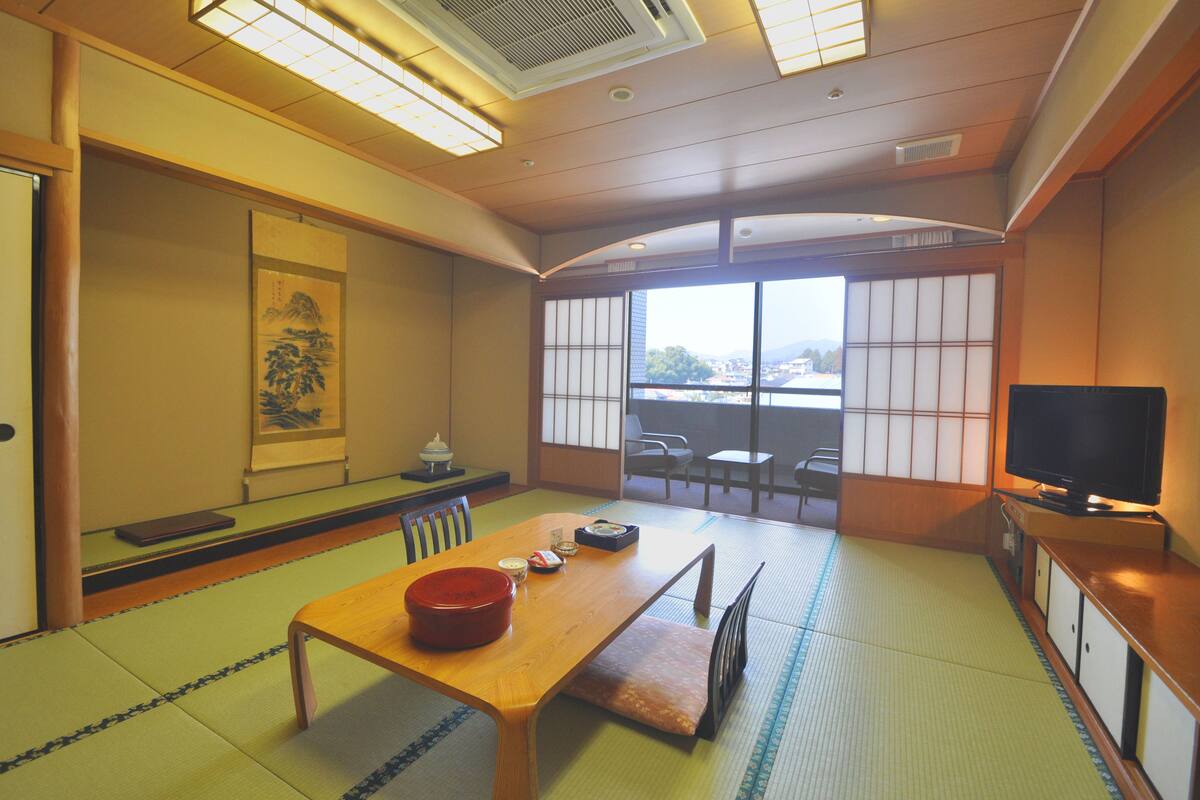 Ett traditionellt japanskt rum med tatamimattor på golvet, ett lågt bord med en röd skål, en tv på ett träställ och utsikt genom shojiskärmar.