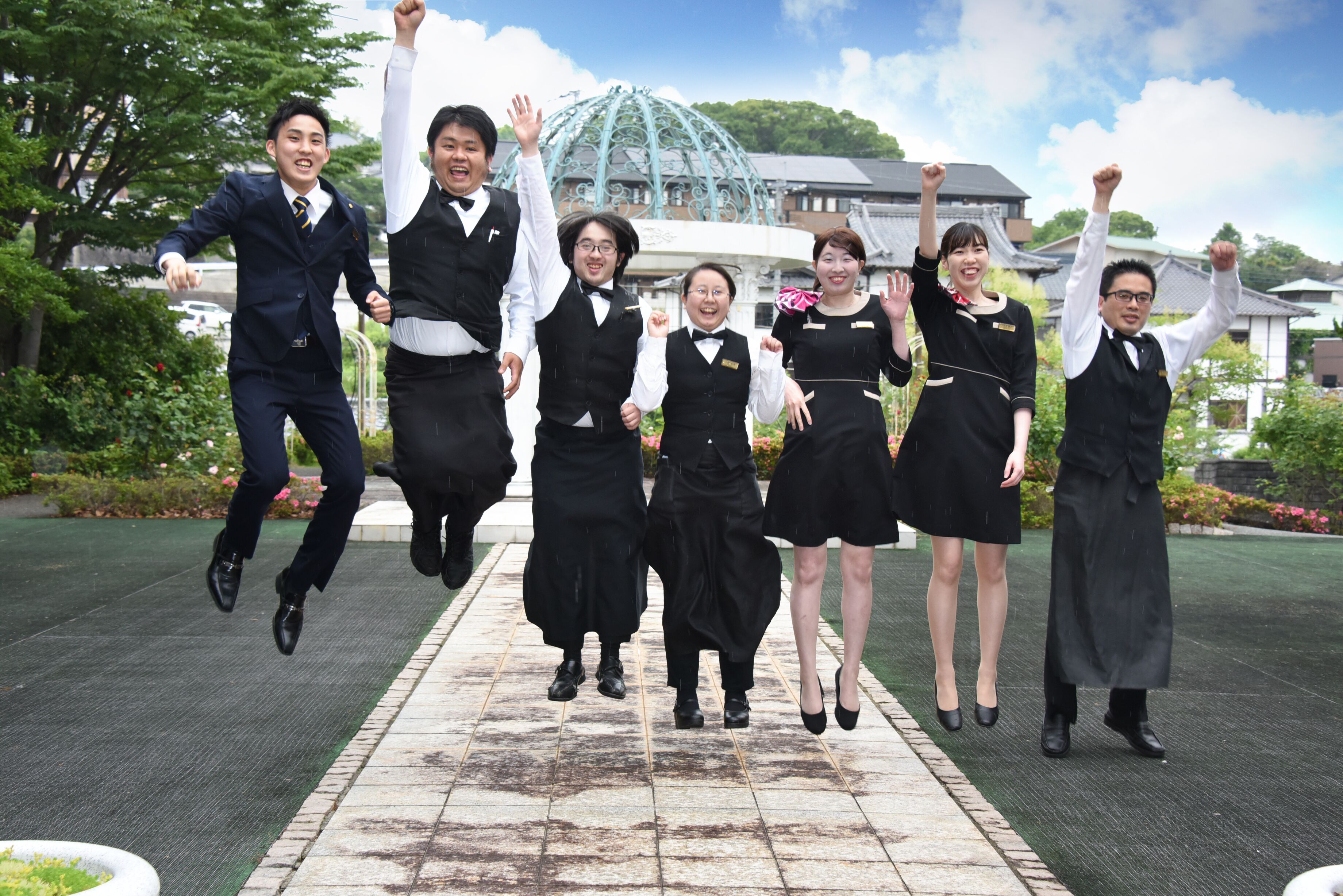 A group of people in formal attire jumping in a garden with a traditional building in the background.