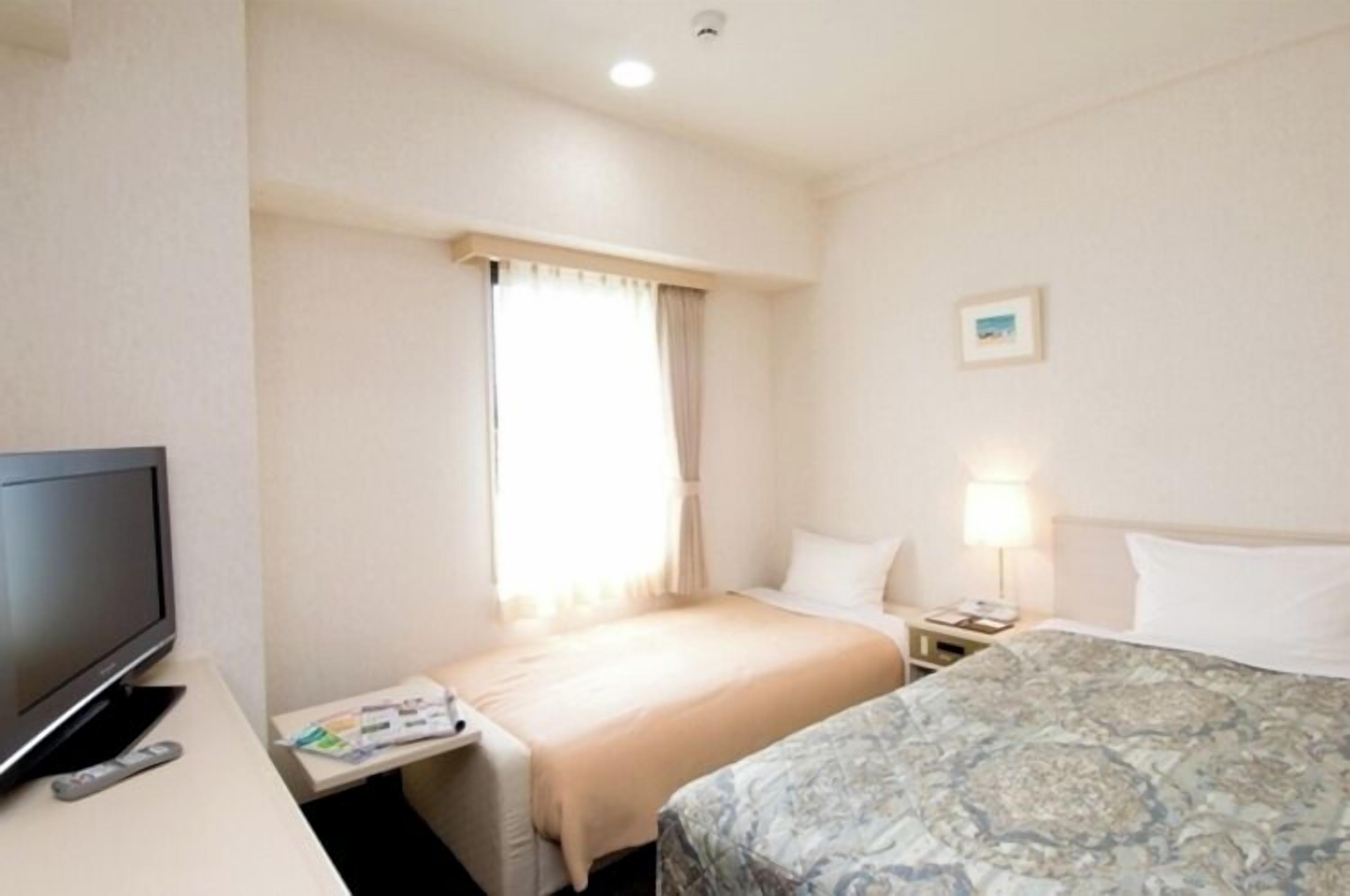 A hotel room with a bed, a TV, a bedside table with a lamp, and a window with curtains.