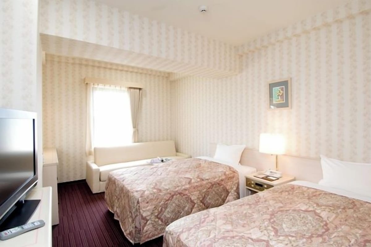 A hotel room with two beds, a TV, a desk, and a window with curtains.