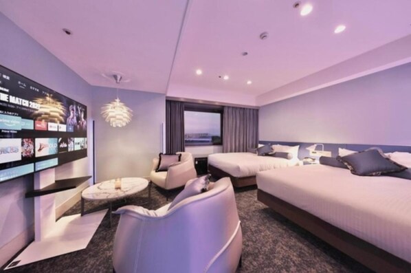A modern hotel room with a large TV screen, a sofa, a bed, and a view of the city.