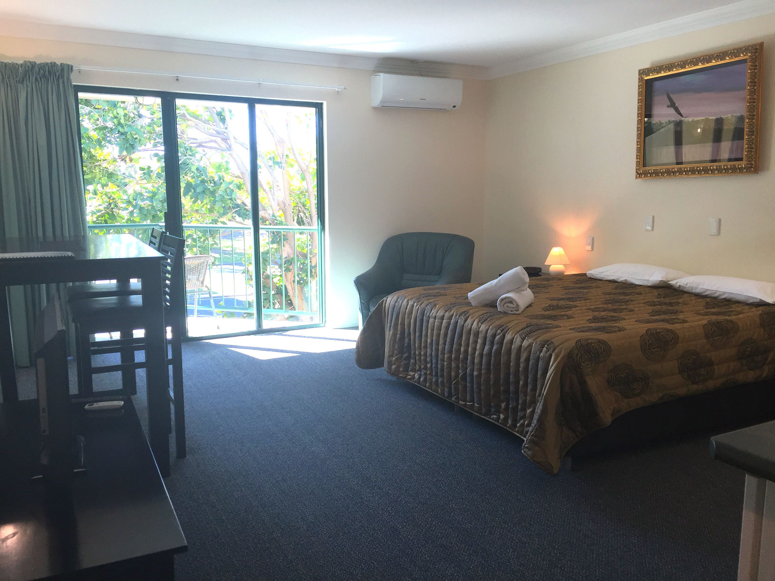 A hotel room with a bed, a desk, a chair, a balcony with a view of greenery, and an air conditioning unit.