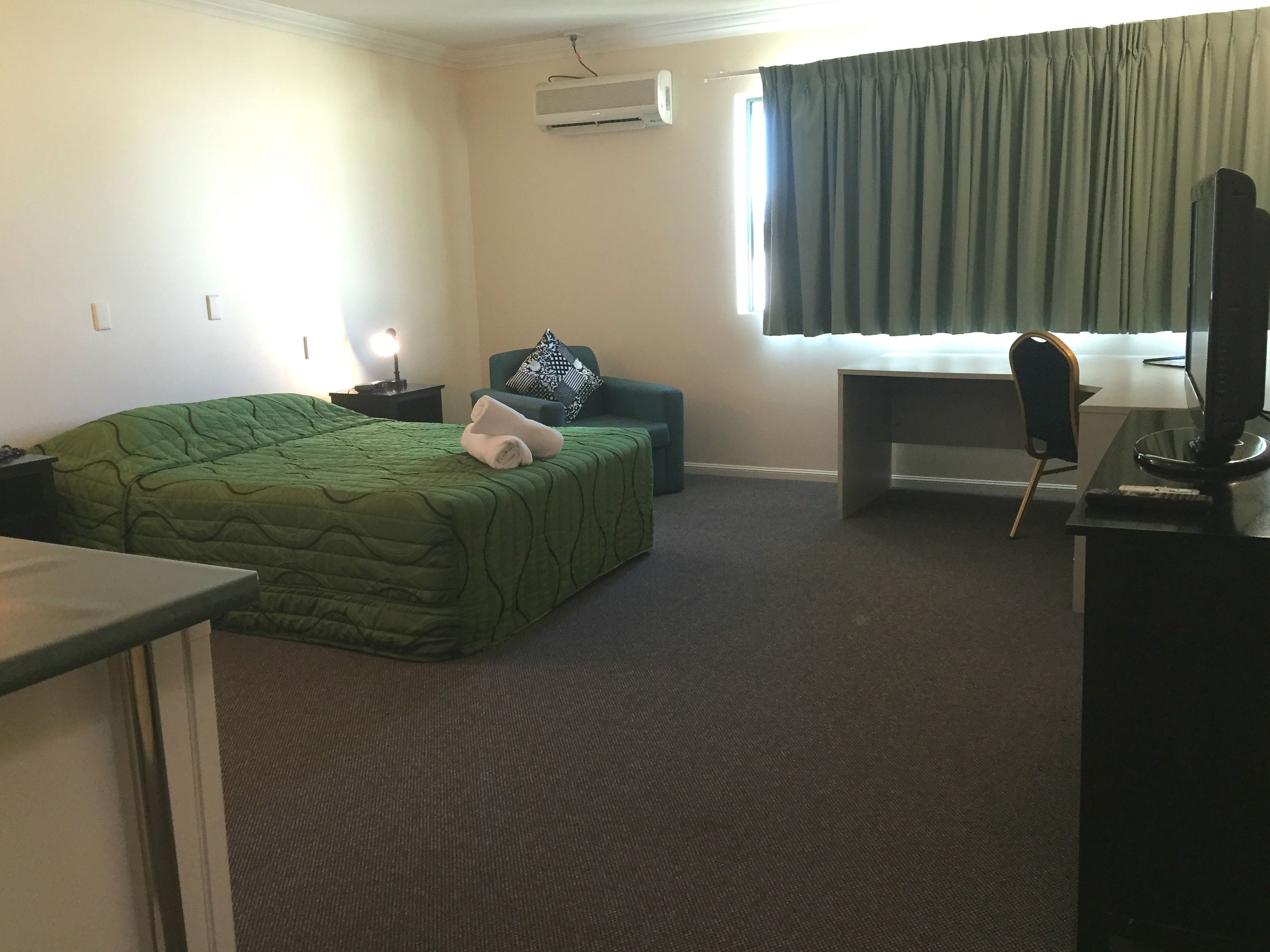 A hotel room with a green bed, a desk with a computer, a chair, a TV, and a window with curtains.