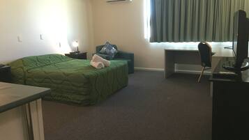 A hotel room with a green bed, a desk with a computer, a chair, a TV, and a window with curtains.