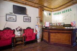 A reception area with a wooden reception desk, a large decorative vase, two red upholstered chairs, and a wall-mounted television. - Green Hotel (Hanoi)