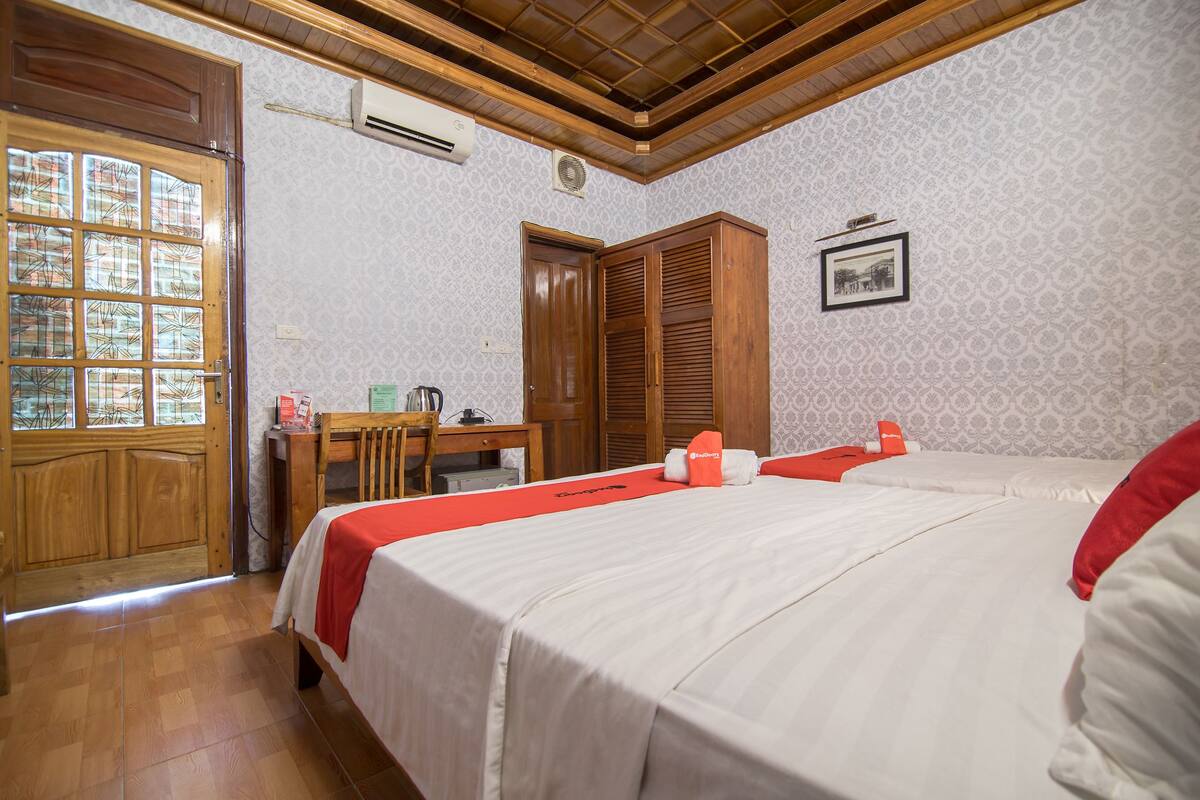 Habitación de hotel con puerta de madera, un escritorio con silla, cama con ropa blanca y detalles rojos, y un papel tapiz estampado.