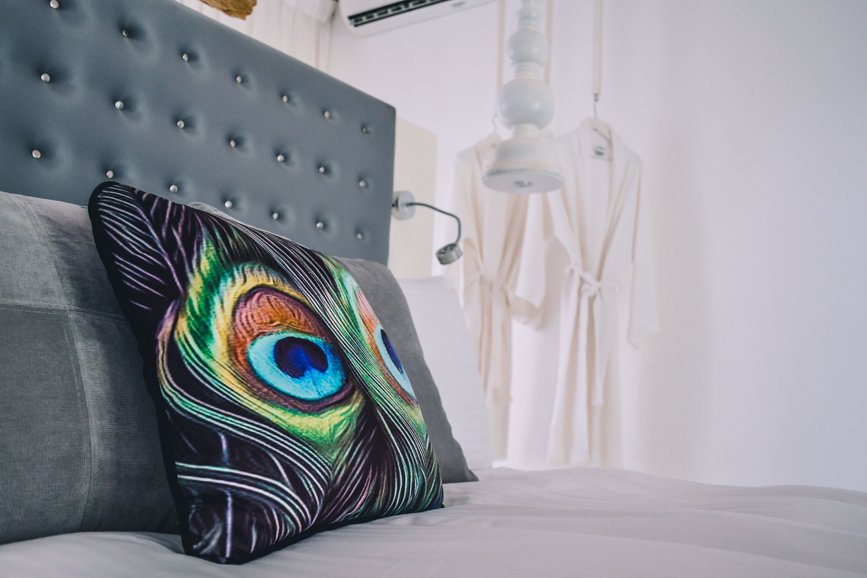 Una cama con cabecera gris y un cojín con una pluma de pavo real de colores.