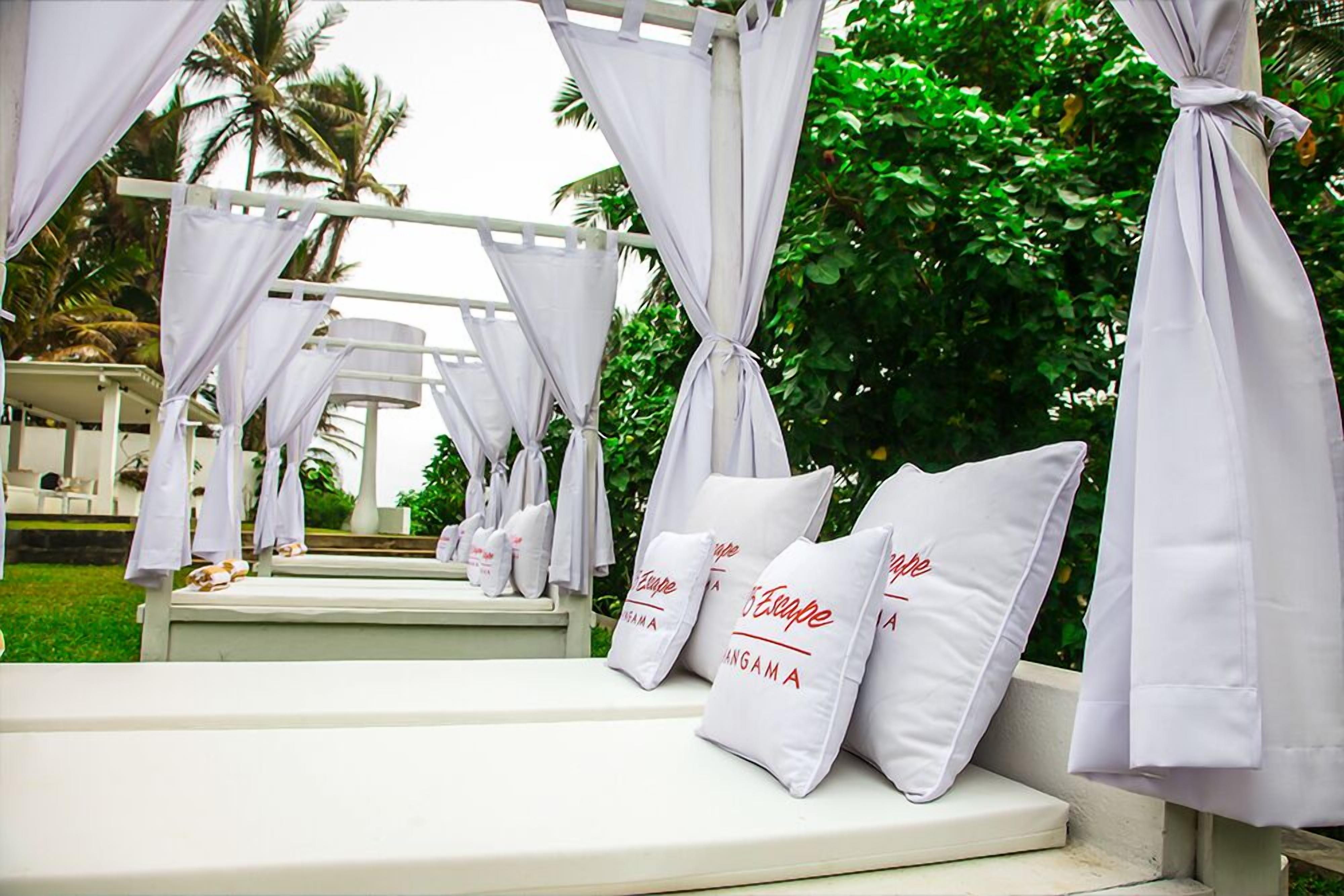 A white outdoor seating area with cushioned benches and white pillows with text, surrounded by greenery and a white structure.