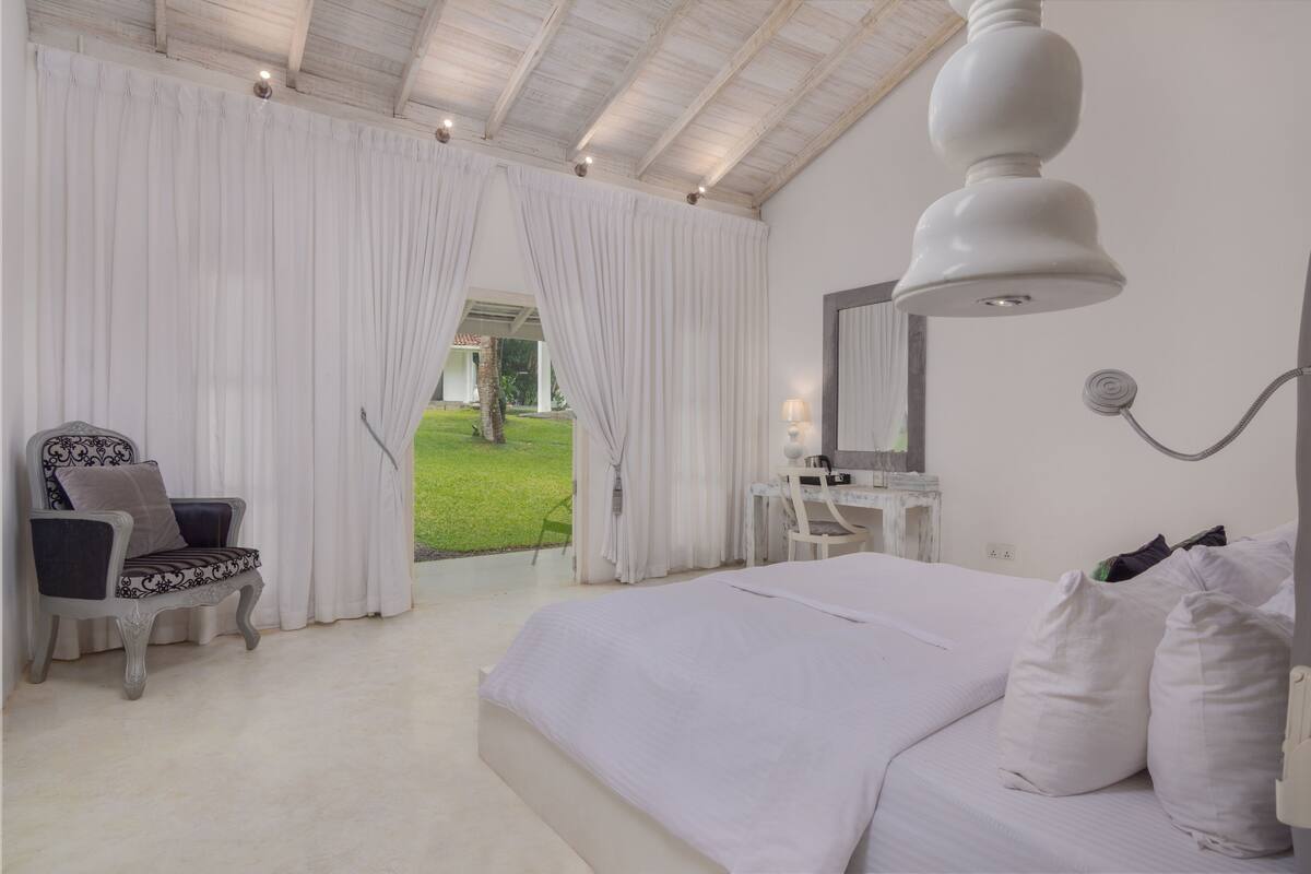 Un dormitorio con una cama grande, una silla, un escritorio y vistas a un jardín a través de una ventana.