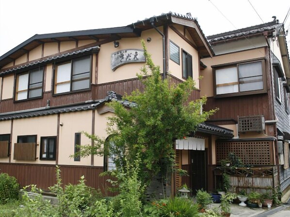 A traditional Japanese house with a wooden exterior, multiple windows, and a small garden area with potted plants. - Shikikaoru Yado Sadae (Kami)