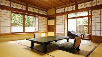 A tatami mat room with a low table and chairs, large windows with shoji screens, and a view of trees outside.