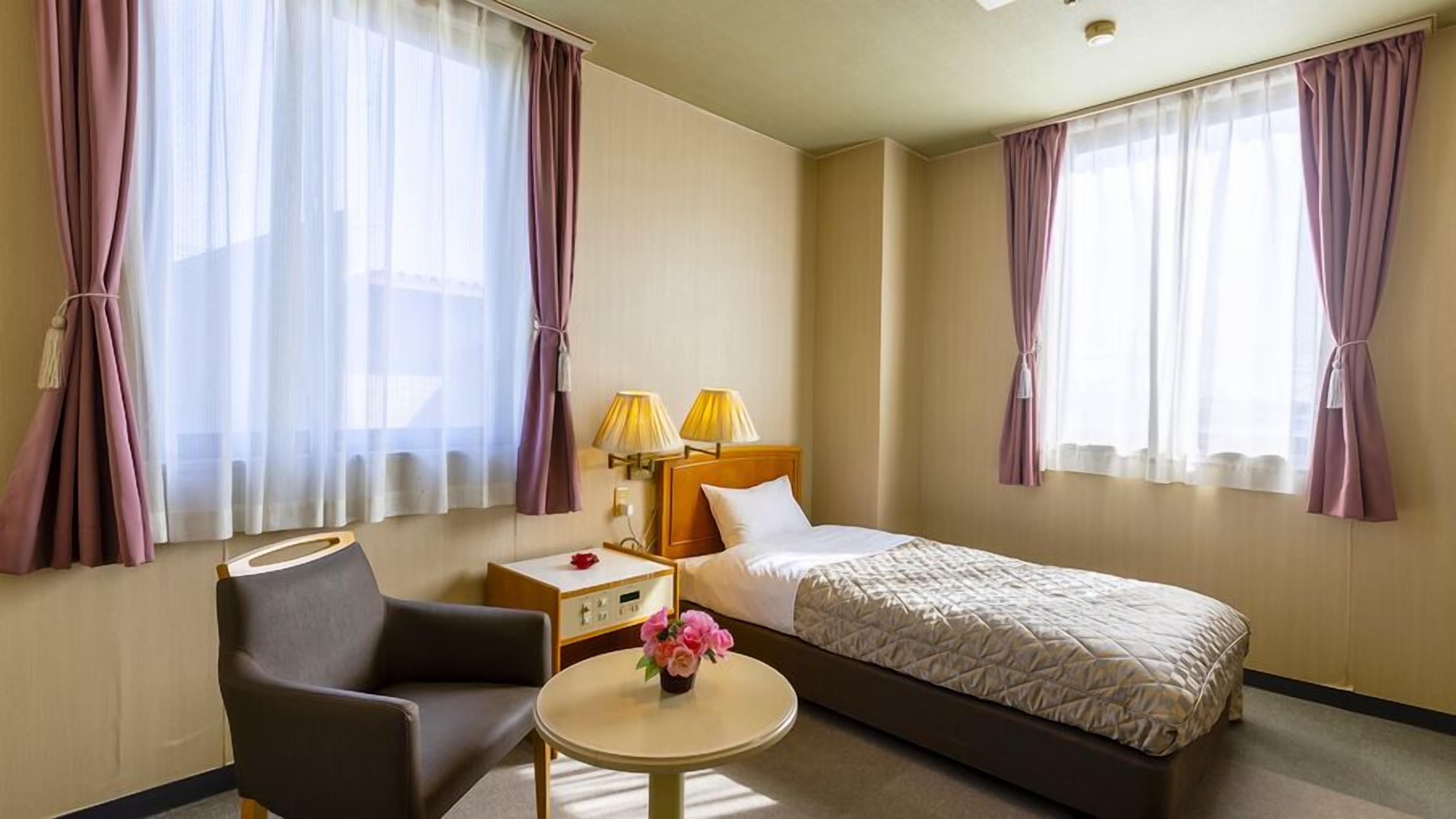Een hotelkamer met een bed, een stoel, een kleine tafel met een bloevas en twee lampen op een nachtkastje.