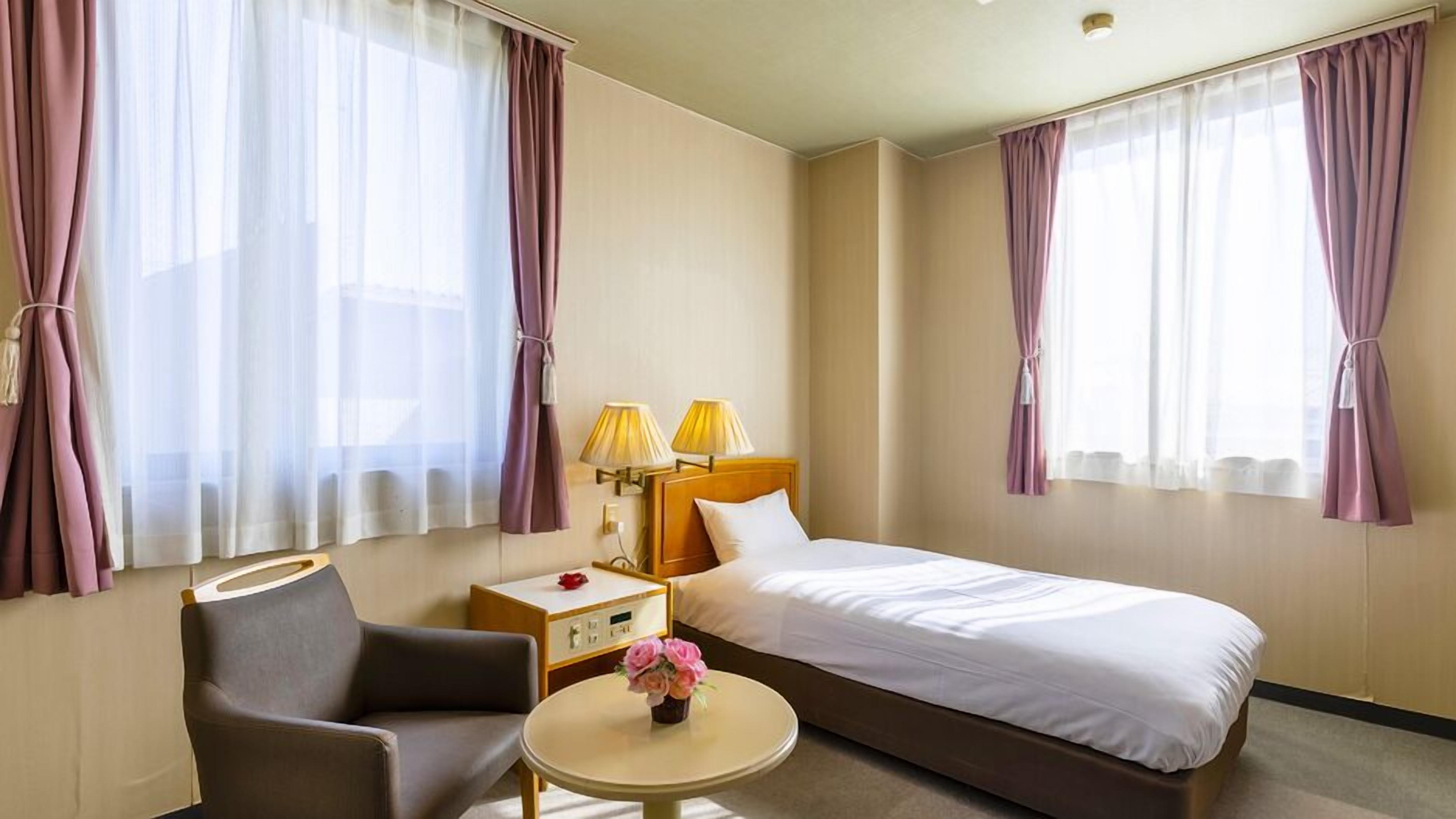 Een hotelkamer met een bed, een stoel, een klein tafeltje met een bloevas en twee lampen op een nachtkastje.