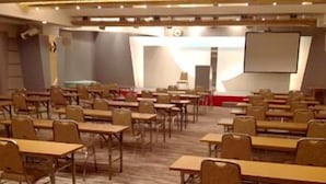 Un salón de conferencias con filas de mesas y sillas, un escenario con una pantalla y suelo alfombrado.
