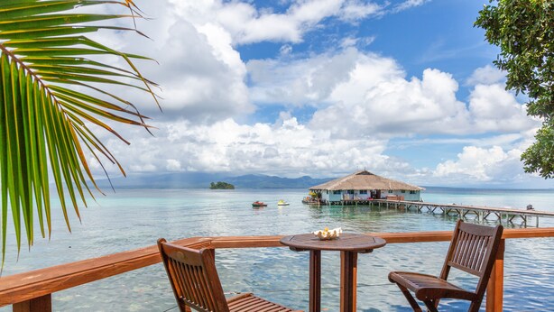 A wooden deck with two chairs and a table overlooking a clear blue sea, a thatched-roof structure on stilts in the distance, and a small island with boats. - Fatboys Resort (Mbambanga Island)