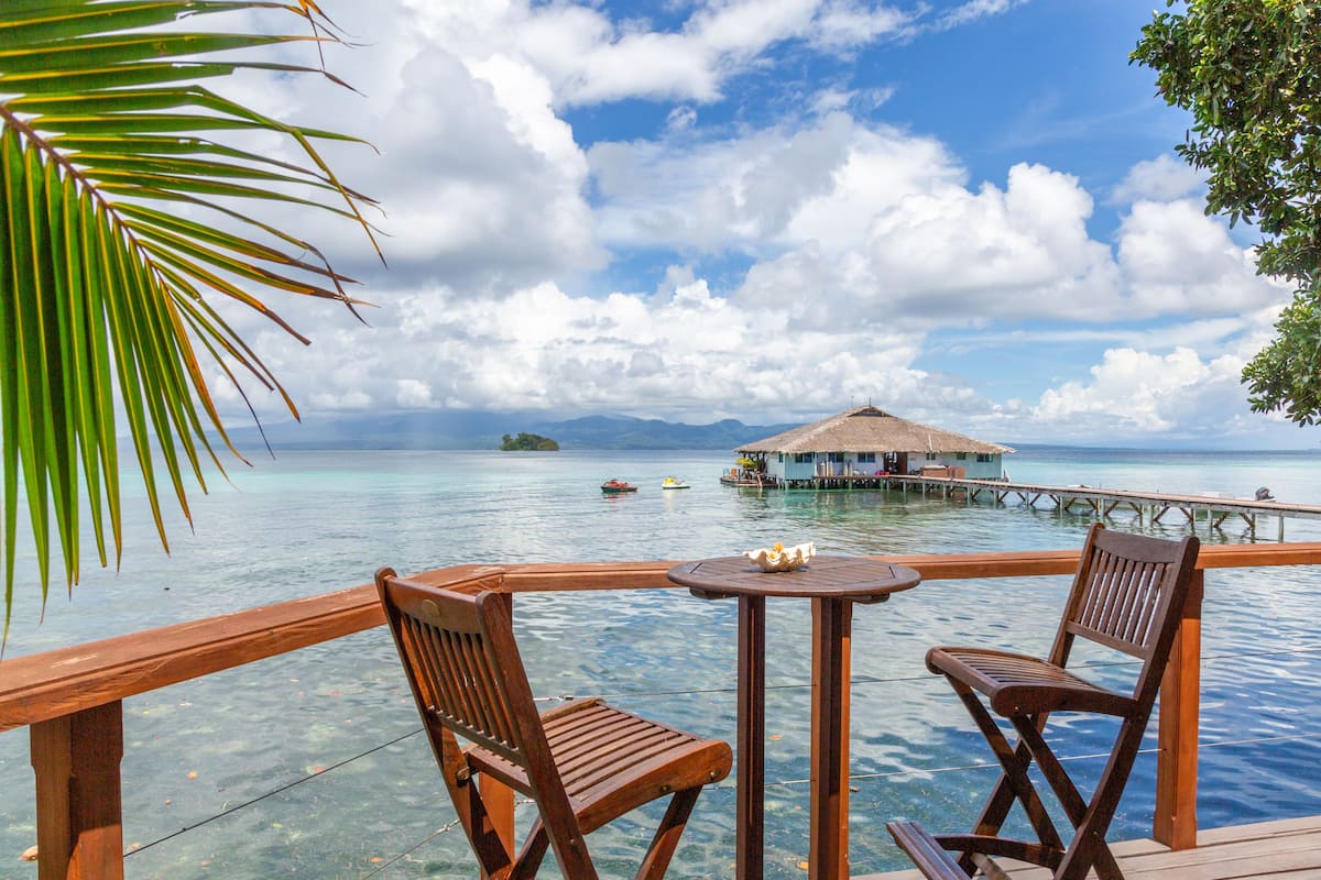 A wooden deck with two chairs and a table overlooking a clear blue sea, a thatched-roof structure on stilts in the distance, and a small island with boats.