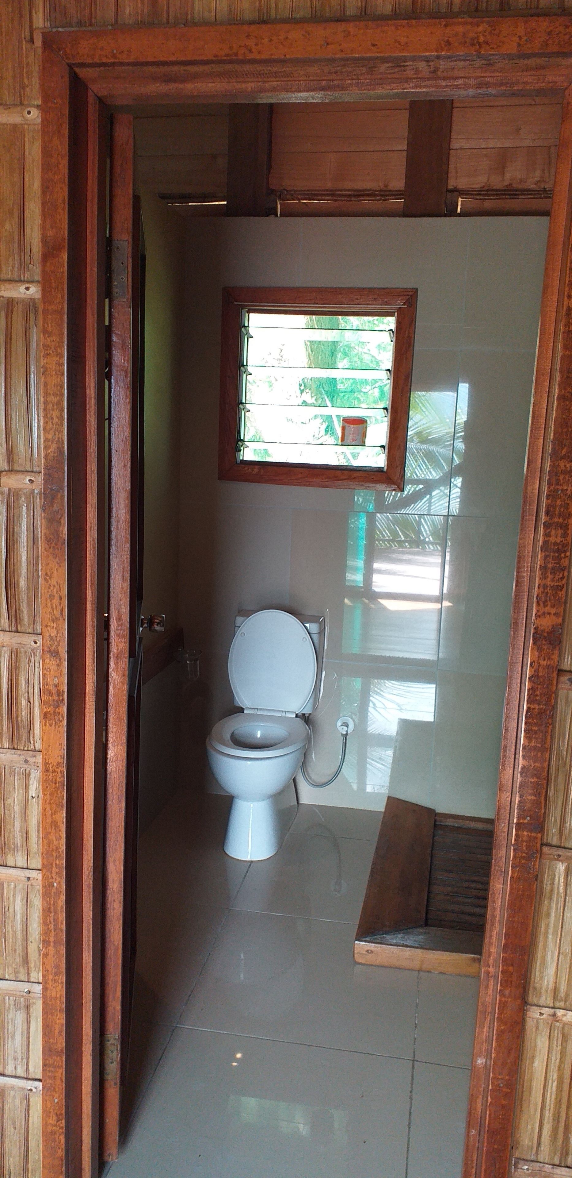 A small, enclosed bathroom with a toilet, a window, and a wooden door frame.