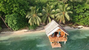 An overwater bungalow with a thatched roof, wooden deck, and a view of the turquoise sea and lush greenery. - Fatboys Resort (Mbambanga Island)