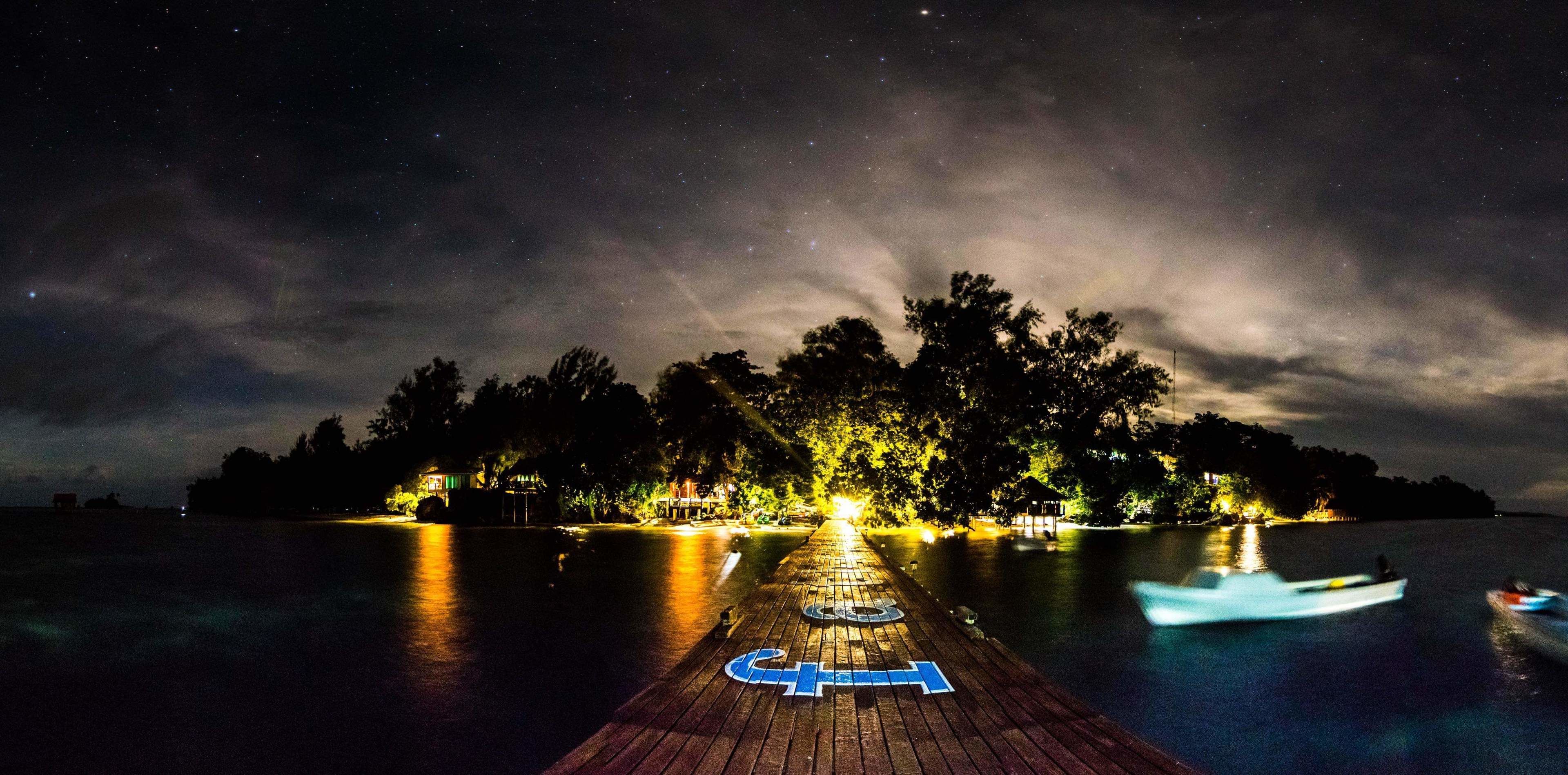 A wooden pier extending into a calm body of water at night, with boats visible and a distant shoreline illuminated by lights.