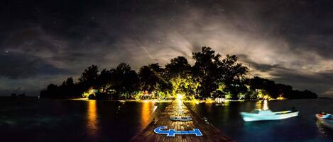 A wooden pier extending into a calm body of water at night, with boats visible and a distant shoreline illuminated by lights.