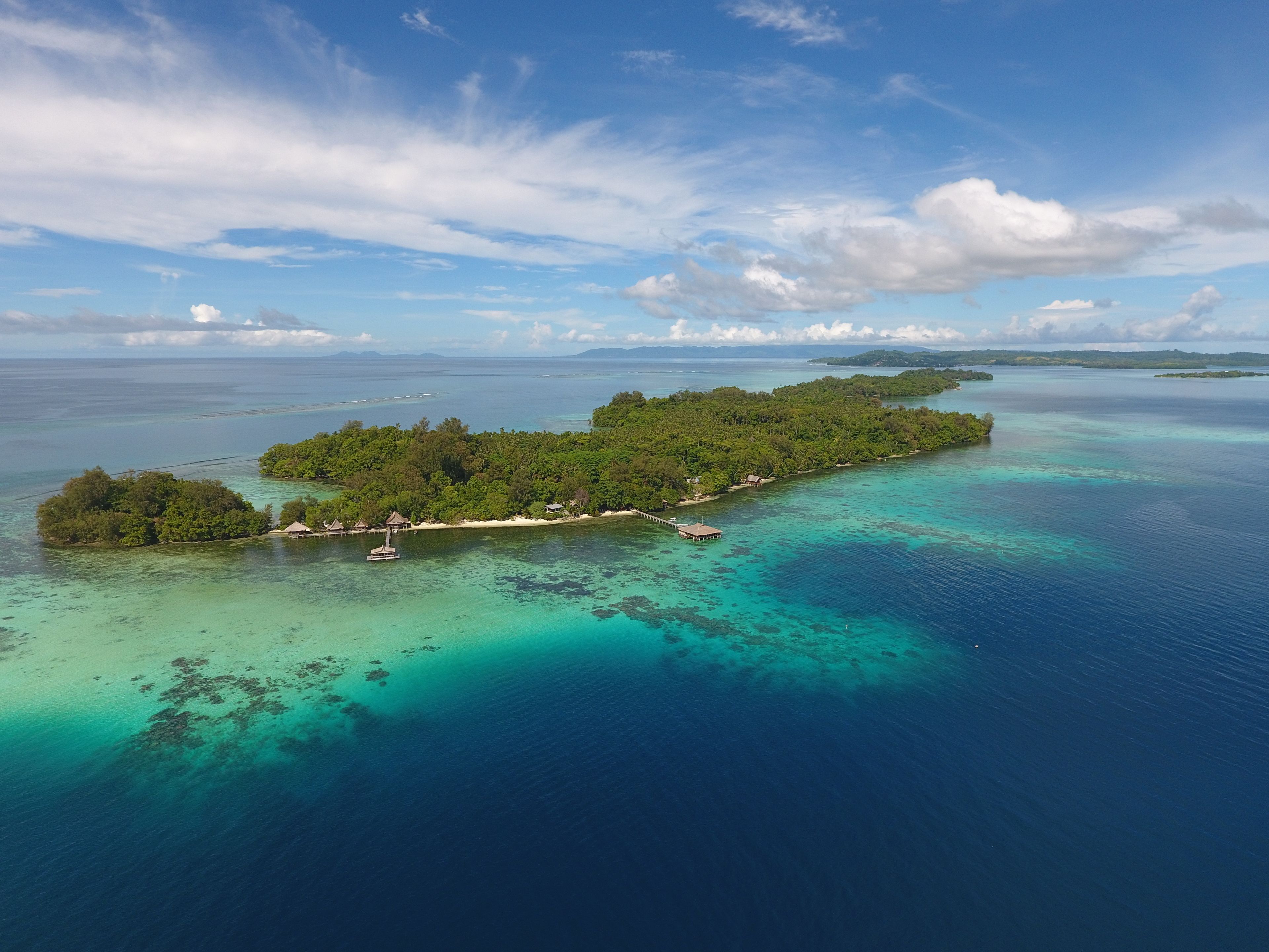 An aerial view of a tropical island with clear turquoise waters, lush greenery, and several small buildings.