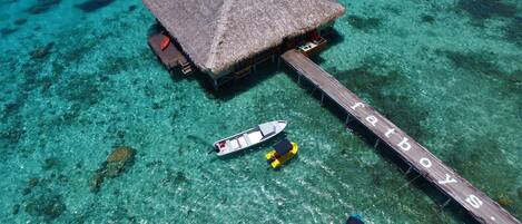 Ein Bungalow auf Stelzen mit einem Reetdach, einer Anlegestelle und klarem türkisblauem Wasser.