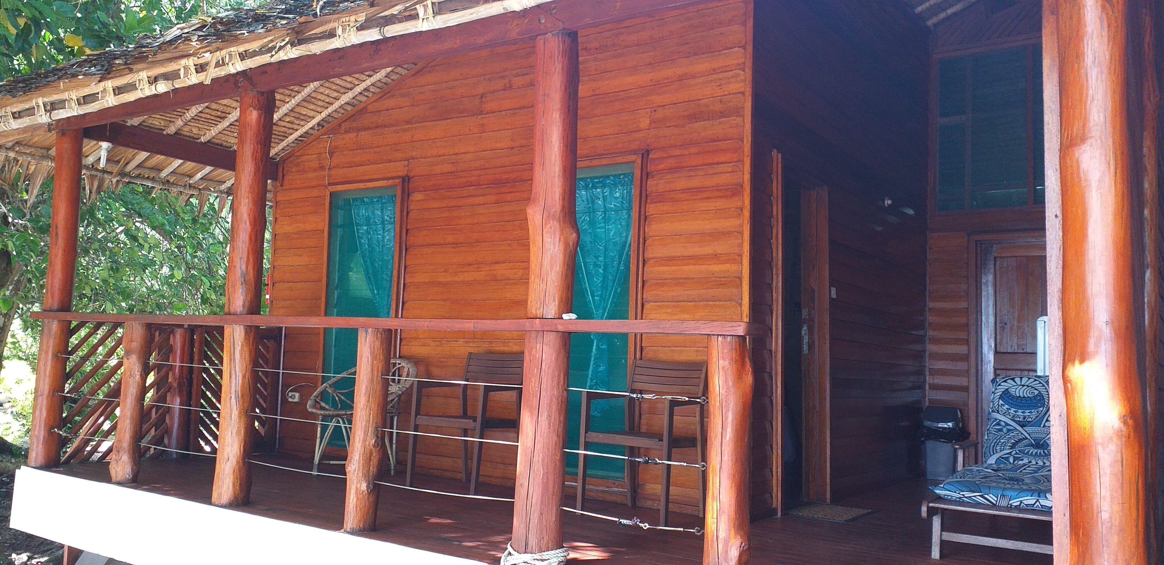A wooden cabin with a porch, two windows with green curtains, and a chair with a patterned cushion.
