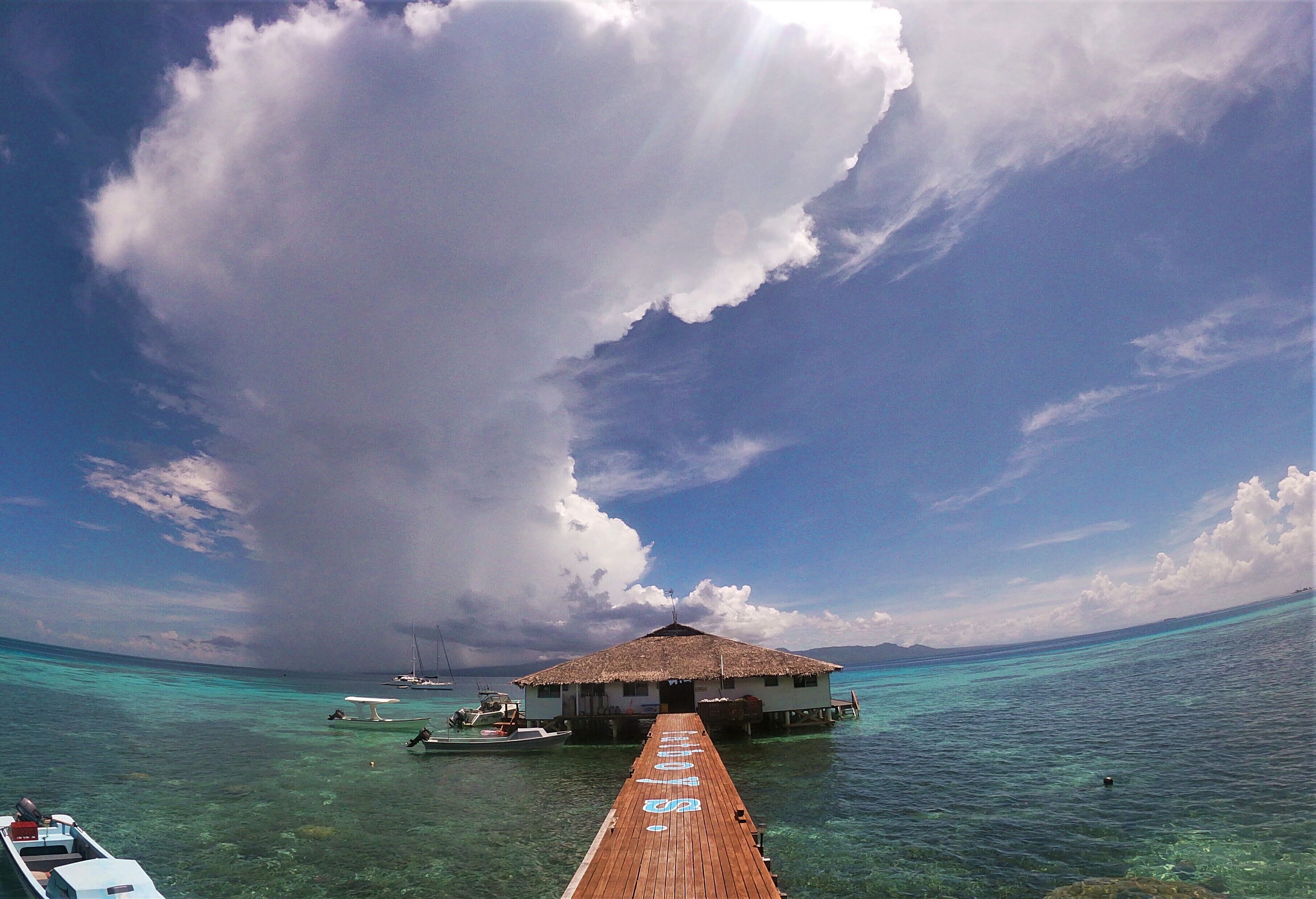 A wooden pier leading to a thatched-roof overwater bungalow with clear turquoise water and a partly cloudy sky.
