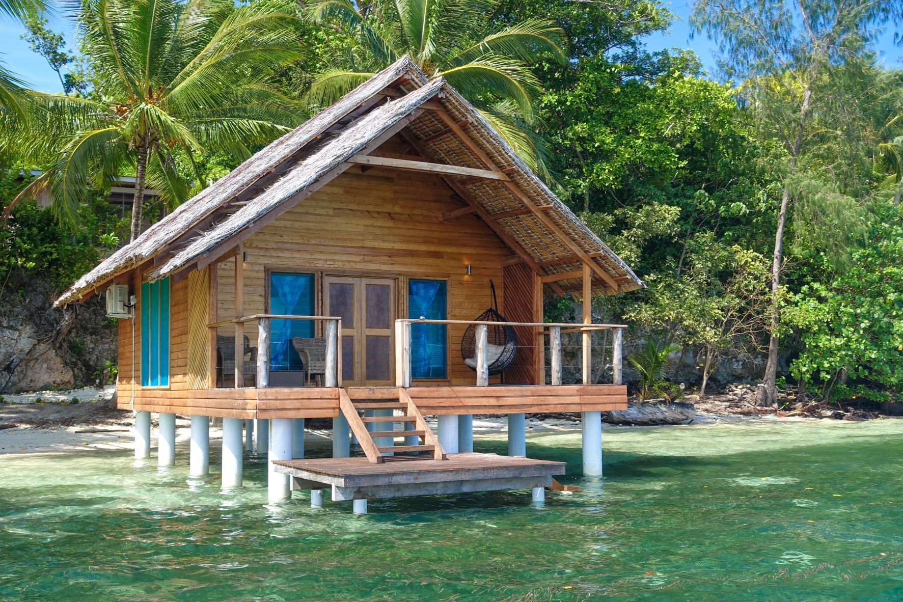 A wooden bungalow with a thatched roof, a balcony, and a view of the surrounding water and greenery.