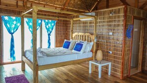 A wooden four-poster bed with white bedding and blue pillows, a small bedside table with a lamp, and a purple rug on a wooden floor. - Fatboys Resort (Mbambanga Island)