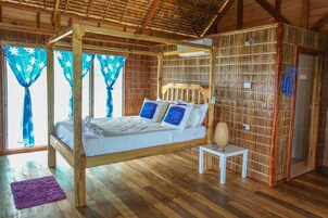 Una cama con dosel de madera, ropa de cama blanca y almohadas azules, una mesita de noche pequeña con una lámpara y un tapete morado sobre un suelo de madera.