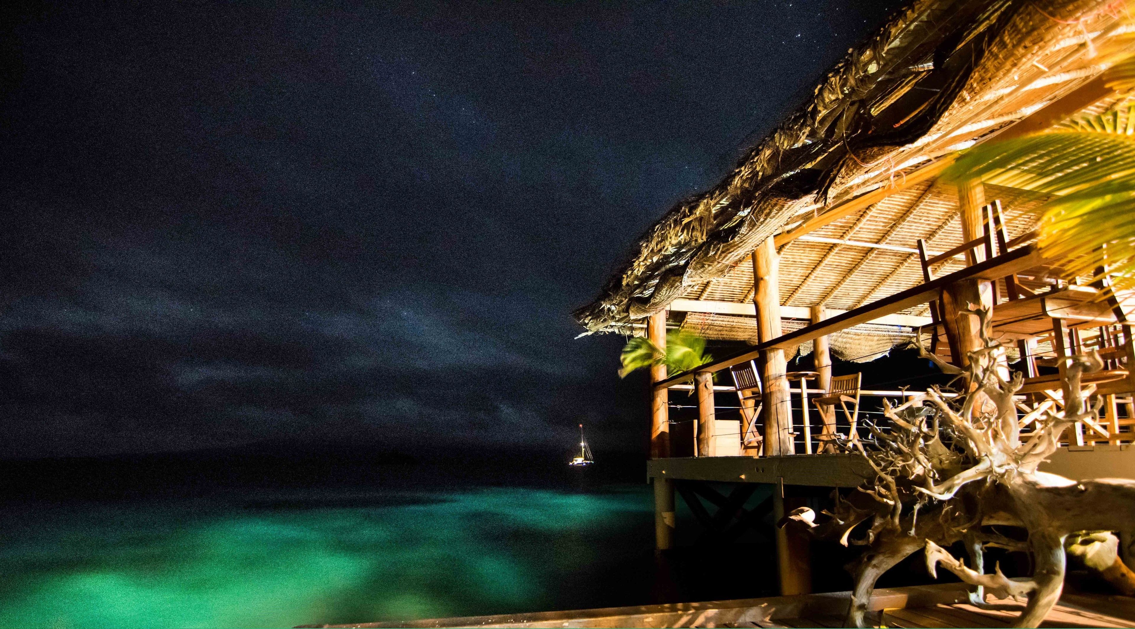 Een rieten dak gebouw met houten leuningen, uitkijkend over turquoise water in de nacht.