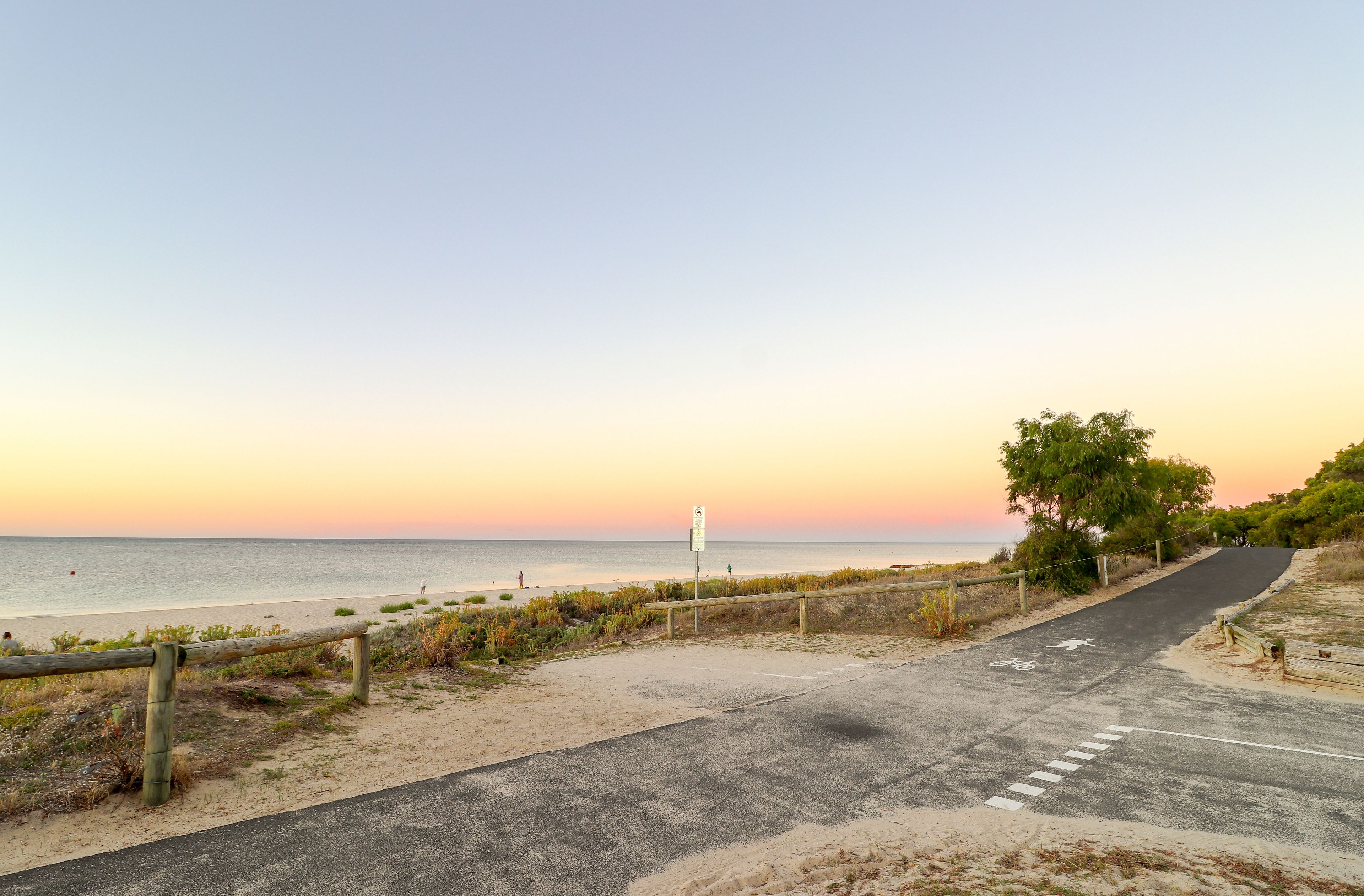Ein gepflasterter Weg führt zu einem Strand, an dem ein Wegweiser und ein Holzzaun stehen.