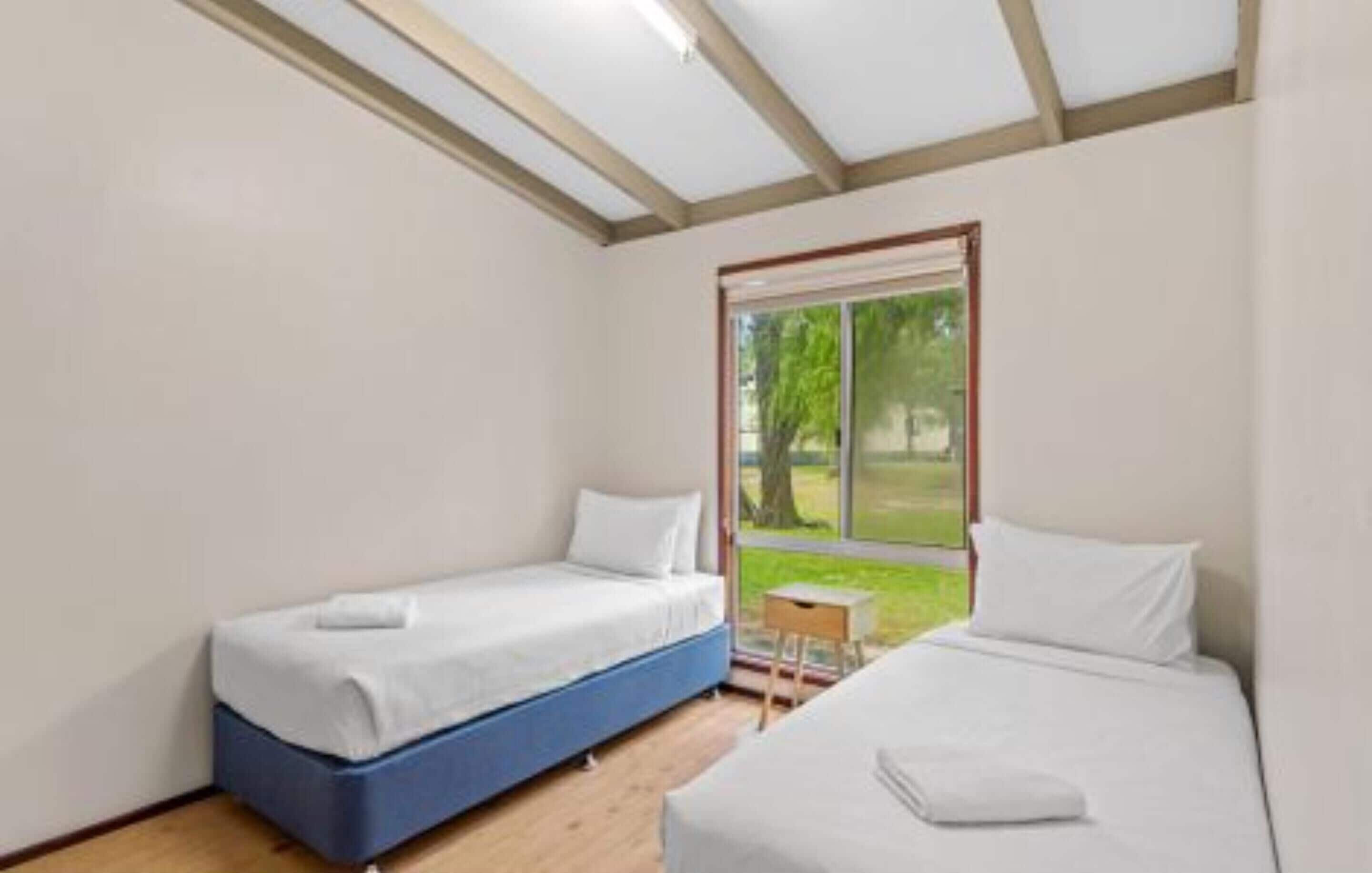 A room with two beds, a small table, and a window with a view of greenery.