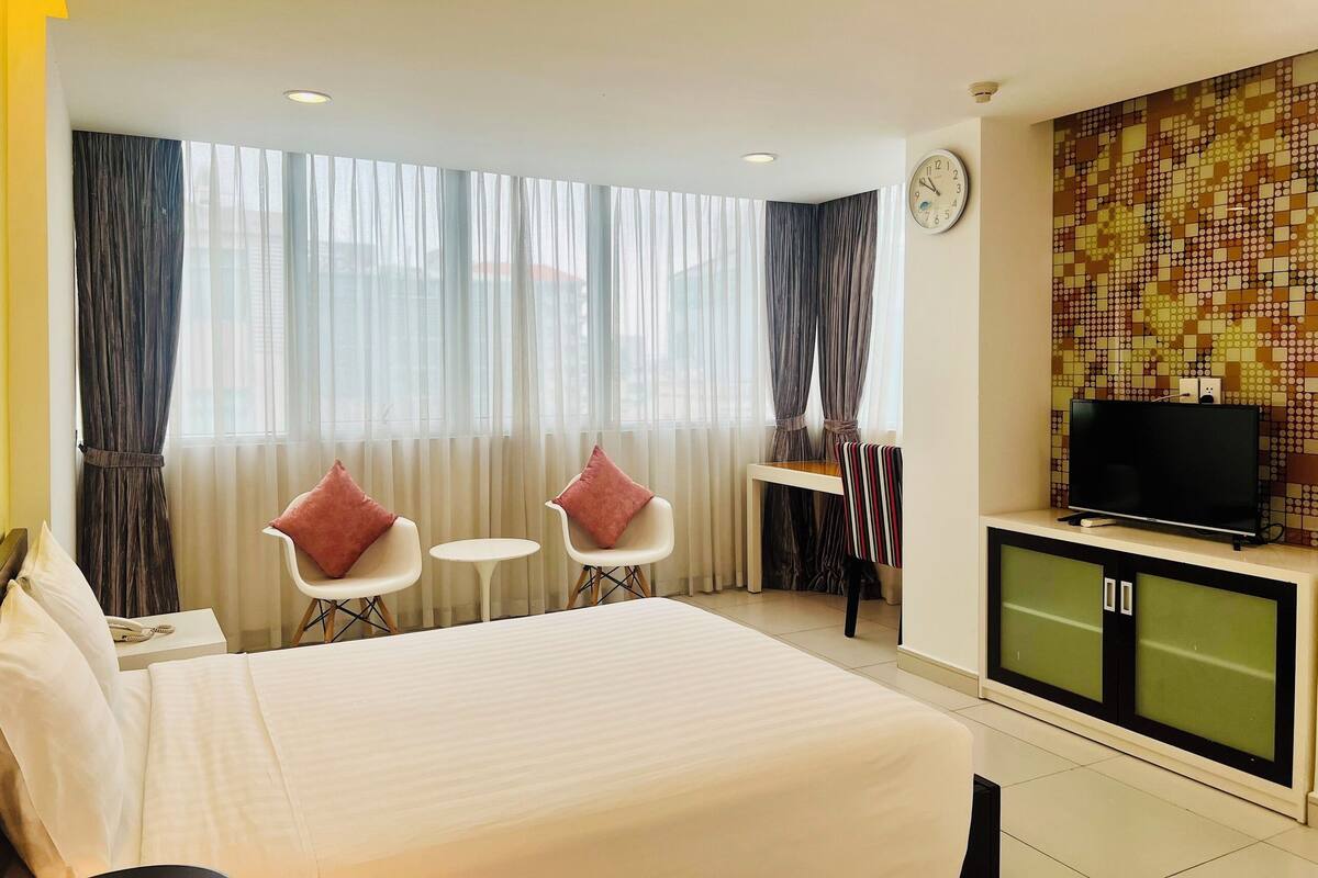 Una habitación de hotel con una cama, dos sillas, una mesita, un televisor y un reloj de pared.