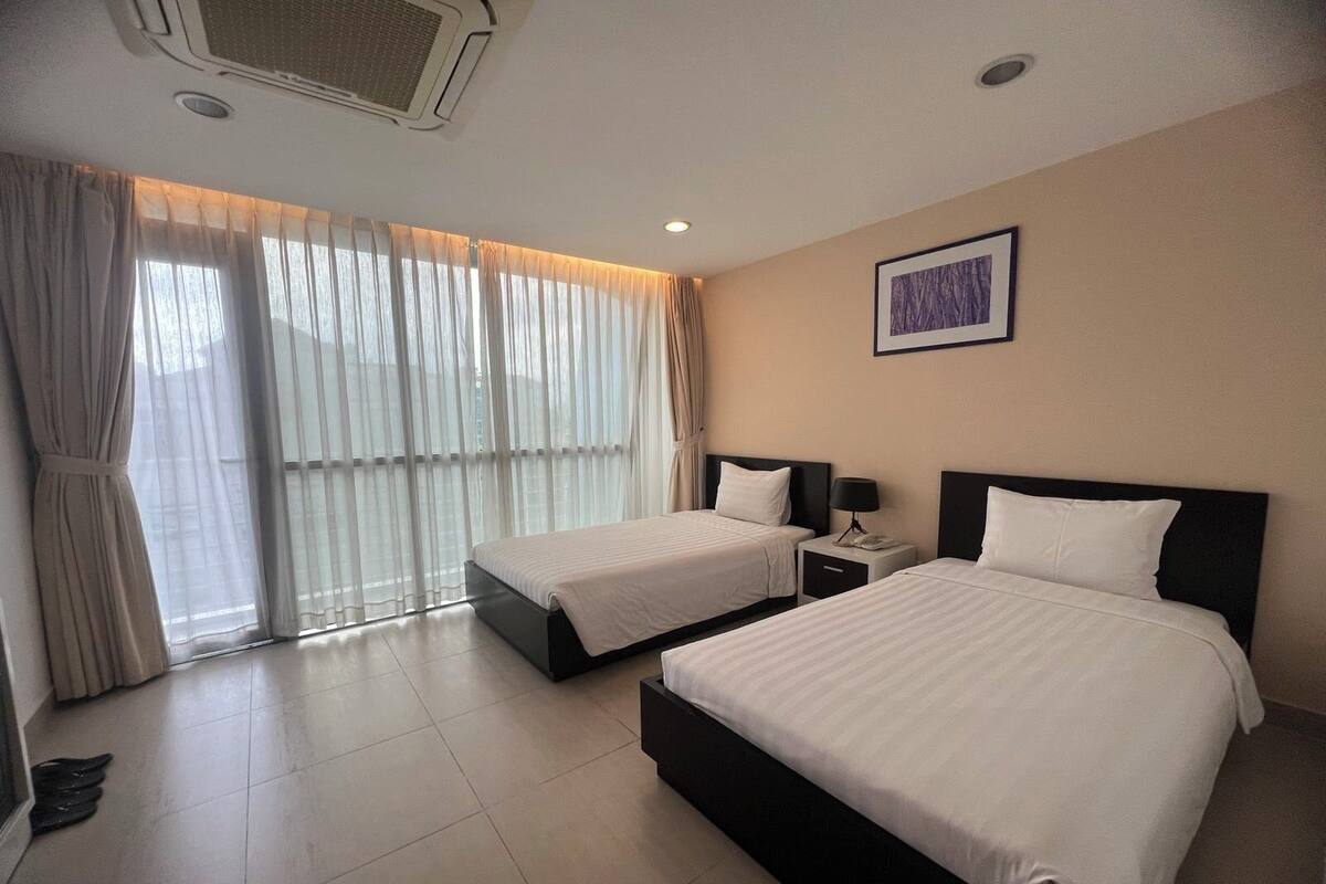 Habitación de hotel con dos camas, un ventanal con cortinas, un cuadro en la pared y un respiradero en el techo.