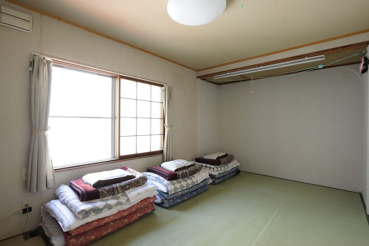A room with tatami flooring, a large window with curtains, and a ceiling fan.